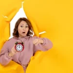 Lady bursting through yellow background pointing at an alarm clock. Shocked look on her face
