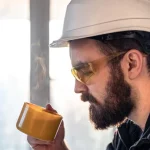 Bearded man in hard hat and goggles drinking a hot drink