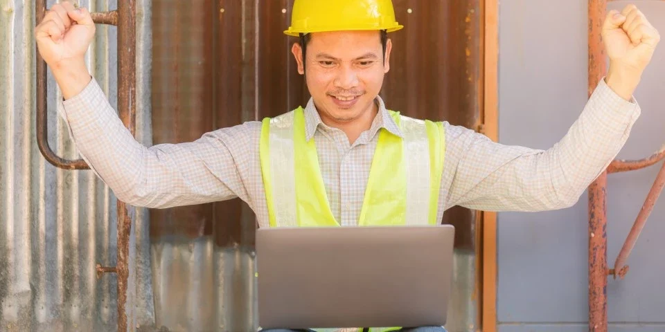 Builder on a building site, with a laptop. He is cheering