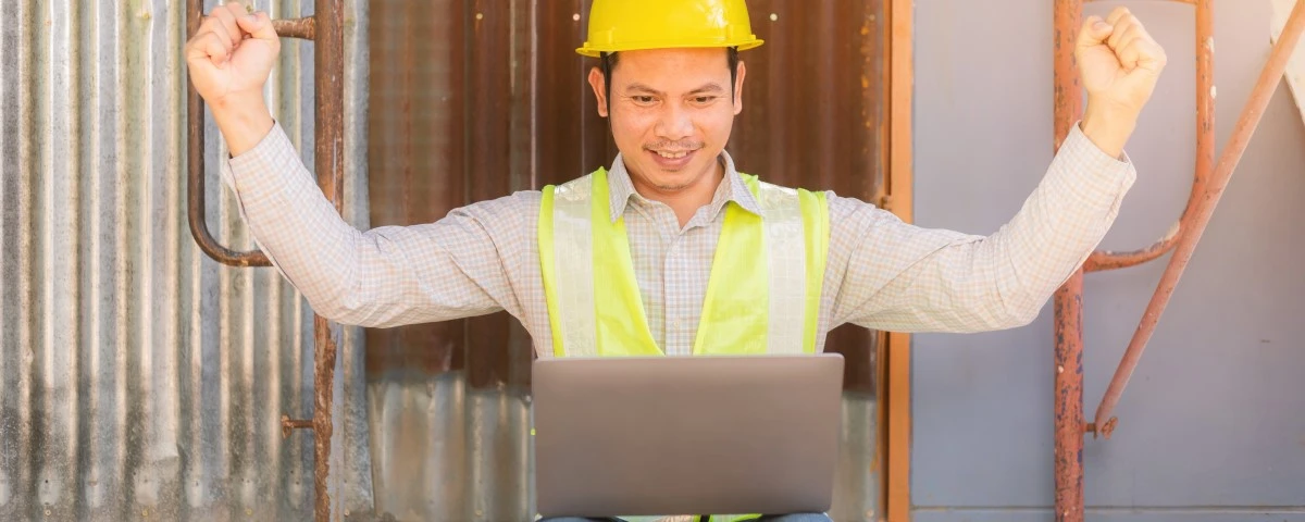 Builder on a building site, with a laptop. He is cheering