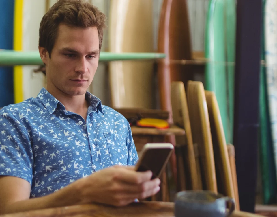 Man sitting at a table scrolling on his phone. He is in front of a display of surfboards.