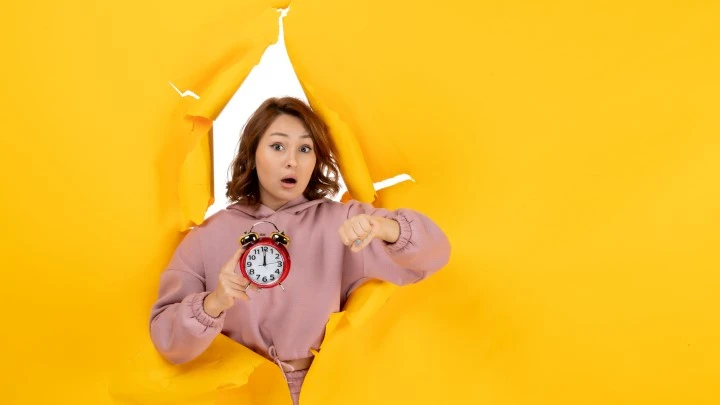 Lady bursting through yellow background pointing at an alarm clock. Shocked look on her face