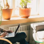 Young tattooed man sitting on a chair with a laptop on his lap. He is looking at the screen. There is a guitar and some headphones on the chair opposite him.