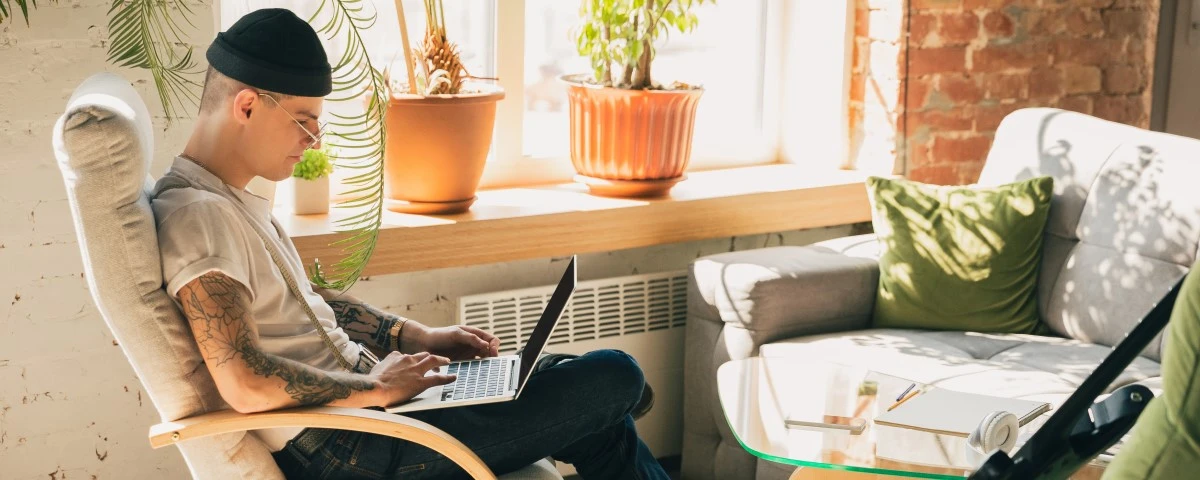 Young tattooed man sitting on a chair with a laptop on his lap. He is looking at the screen. There is a guitar and some headphones on the chair opposite him.