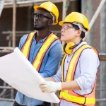 Two men at a construction site, holding a floor plan and looking up