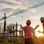 Two builders in hi-vis vests and hard hats observe a building site. the one on the left is pointing at a crane