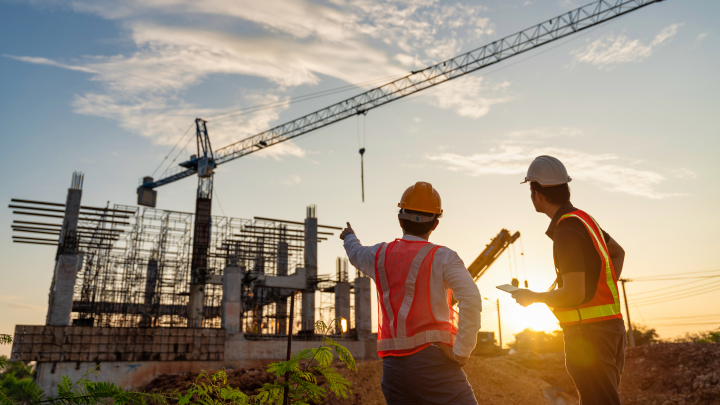 Two builders in hi-vis vests and hard hats observe a building site. the one on the left is pointing at a crane