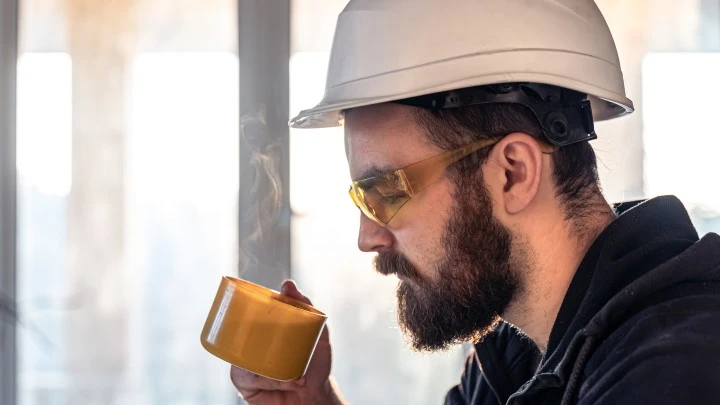 Bearded man in hard hat and goggles drinking a hot drink