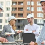 Builders in suits talking, whilst one is on his laptop