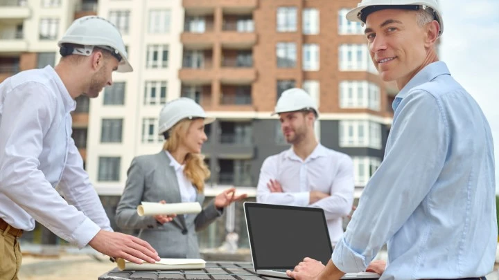 Builders in suits talking, whilst one is on his laptop