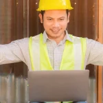 Builder on a building site, with a laptop. He is cheering