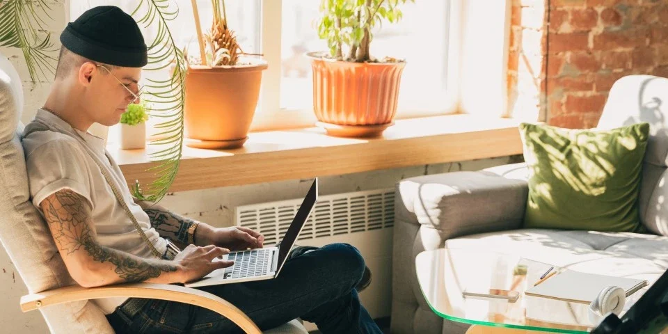 Young tattooed man sitting on a chair with a laptop on his lap. He is looking at the screen. There is a guitar and some headphones on the chair opposite him.